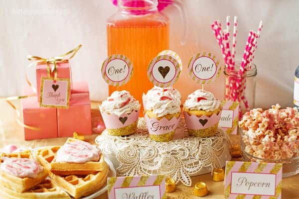 baby girl first birthday pink and gold party decorations table with recipes for waffles, pink cupcakes , popcorn.