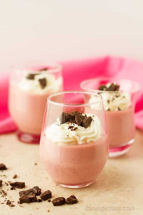 A photo of three glasses with strawberry panna cotta, topped with whipped cream and chocolate. With pink material behind glasses.