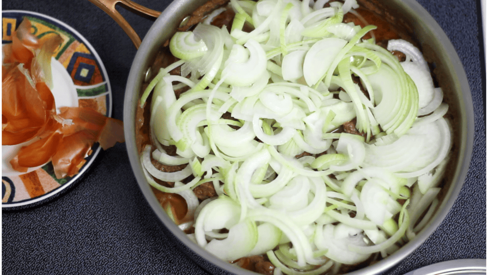Sliced onions on top of the meat in frying pan to make Polish gulasz.